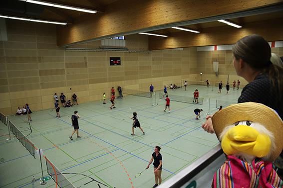 Blick von oben auf ein Badmintonspiel mit mehreren Spielern in einer Sporthalle, ein Pl&uuml;schfrosch in Hut im Vordergrund.