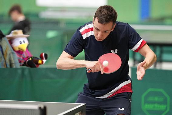 Spieler schl&auml;gt einen Tischtennisball mit einem Schl&auml;ger an einem Tischtennistisch, im Hintergrund steht ein Pl&uuml;sch-Tier.