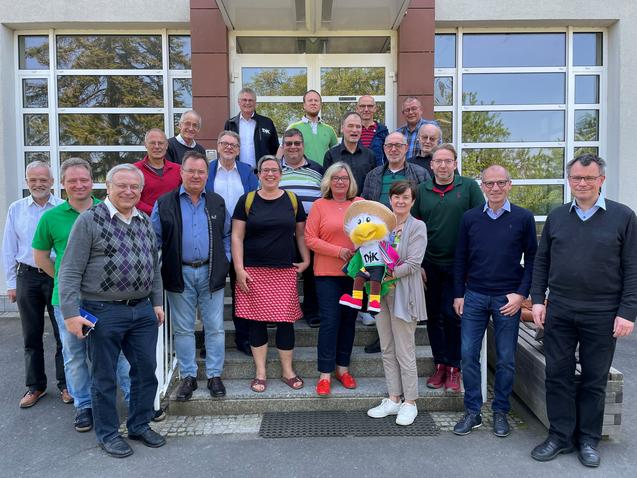 Gruppenfoto von 26 Personen vor einem Gebäude, darunter ein Maskottchen mit buntem Outfit.