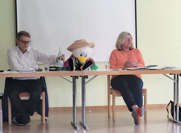 Zwei Personen sitzen an einem Tisch, zwischen ihnen ein Plüschtern mit Hut. Präsentationswand im Hintergrund.