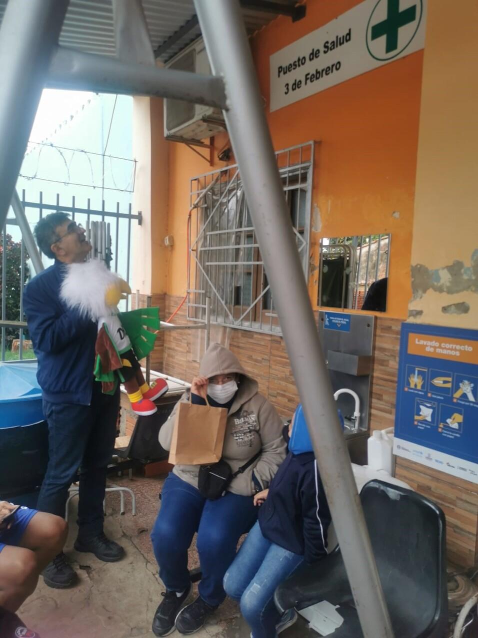 Personen in einem Warteraum vor gesundheitlicher Einrichtung mit Hinweisschild, einer Person h&auml;lt ein Spielzeug.