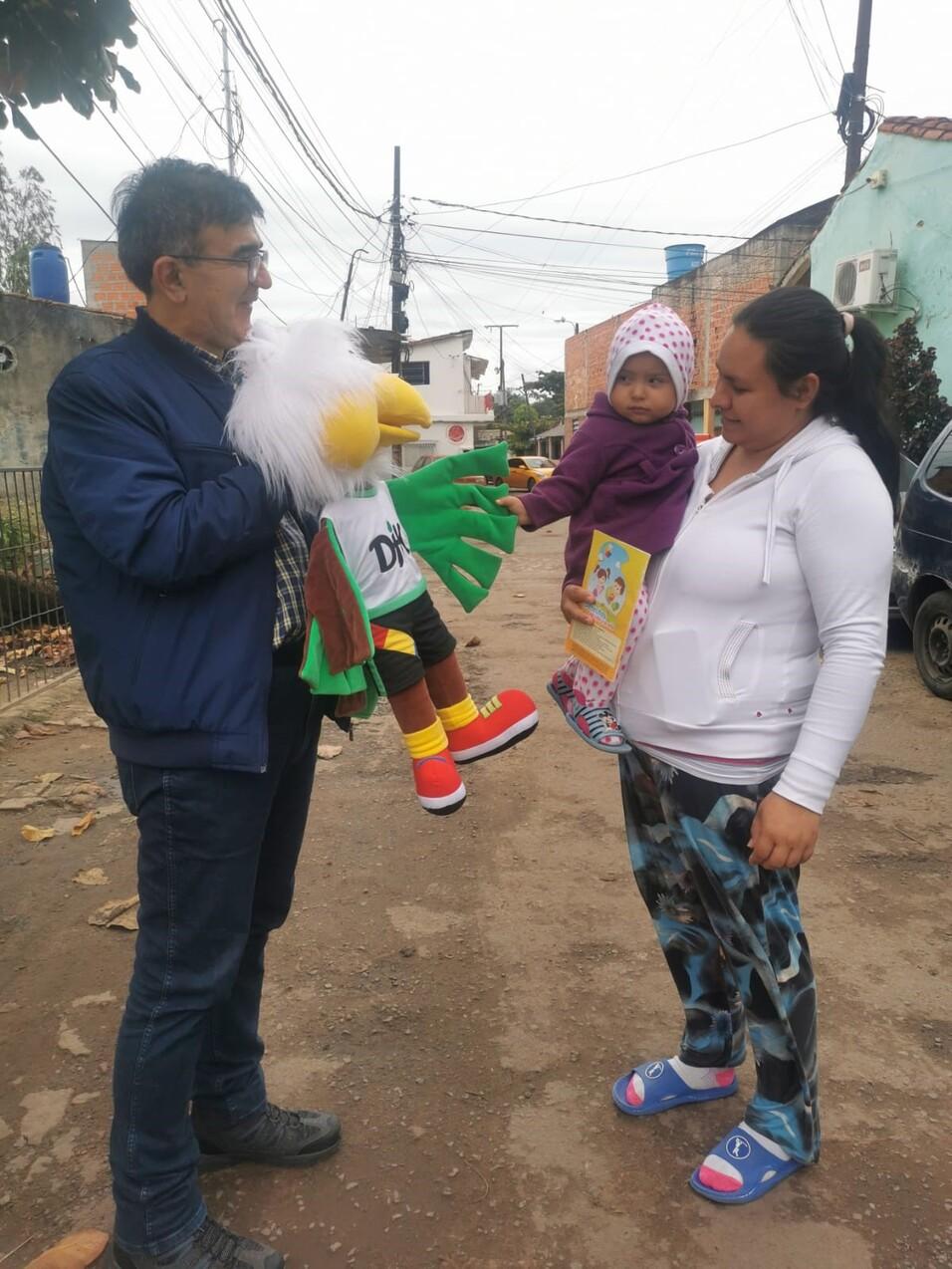 Mann mit Handpuppe und Kind in lila Kleidung steht gegen&uuml;ber einer Frau mit Buch auf einer Stra&szlig;e mit Wohnh&auml;usern.