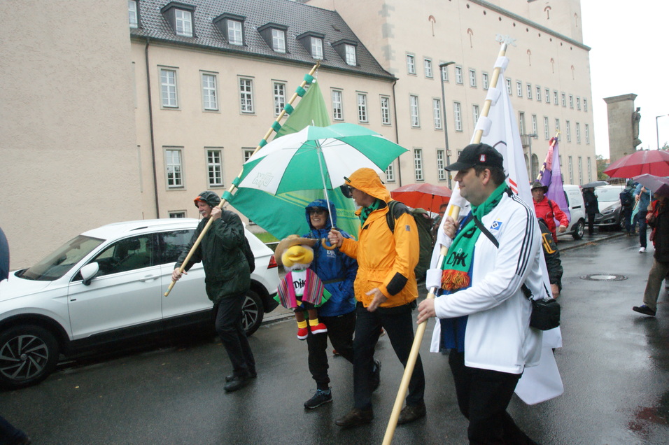 Gruppierung von Menschen bei Regen mit Schirmen und Fahnen, darunter ein bemannter Pl&uuml;schfigur aus einem Umzug.