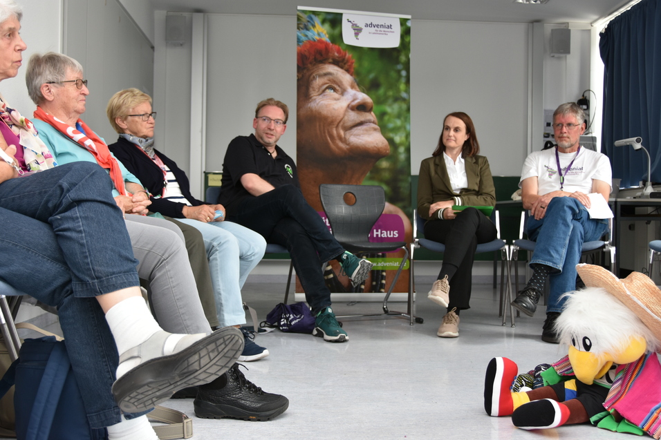 Gruppe von Personen in einem Gespr&auml;ch, umgeben von St&uuml;hlen, mit buntem Plakat im Hintergrund und einem Spielzeug auf dem Boden.