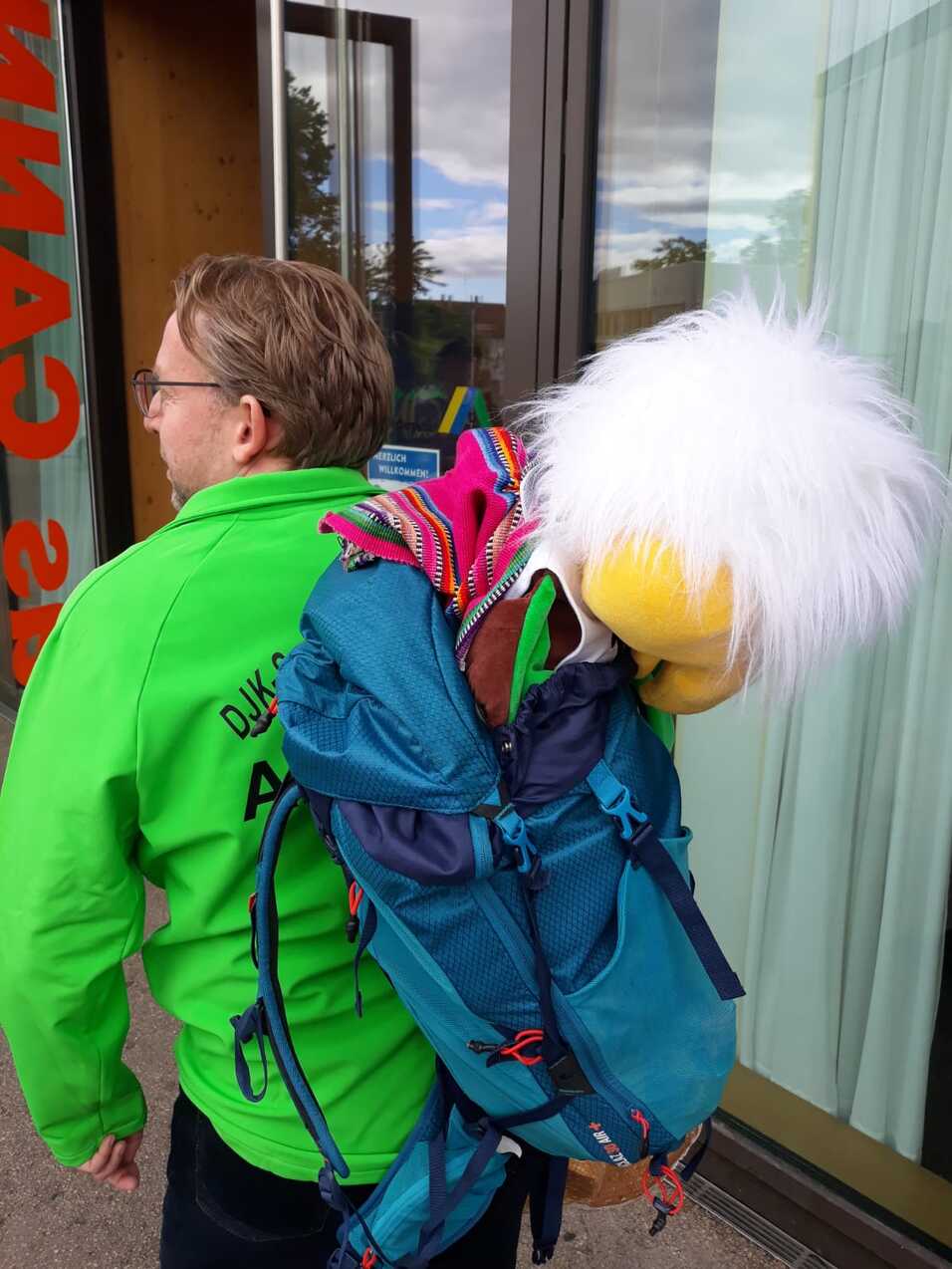 Mann mit Brille tr&auml;gt einen Rucksack, aus dem eine Puppe mit wei&szlig;er Frisur schaut. Im Hintergrund Fenster sichtbar.