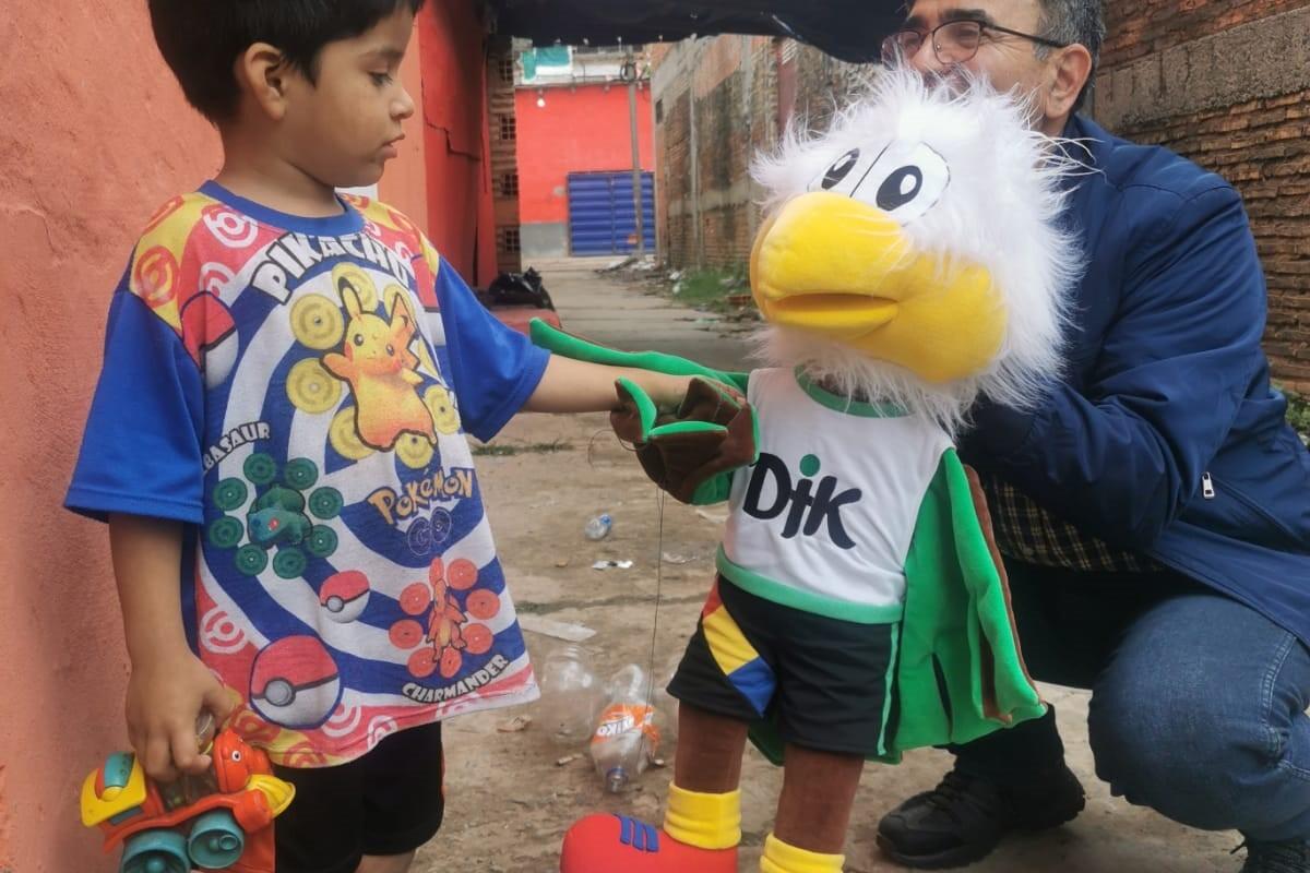 Junge mit Spielzeugauto steht neben einem Mann, der eine gro&szlig;e, l&auml;chelnde Stoffente mit buntem Outfit h&auml;lt.