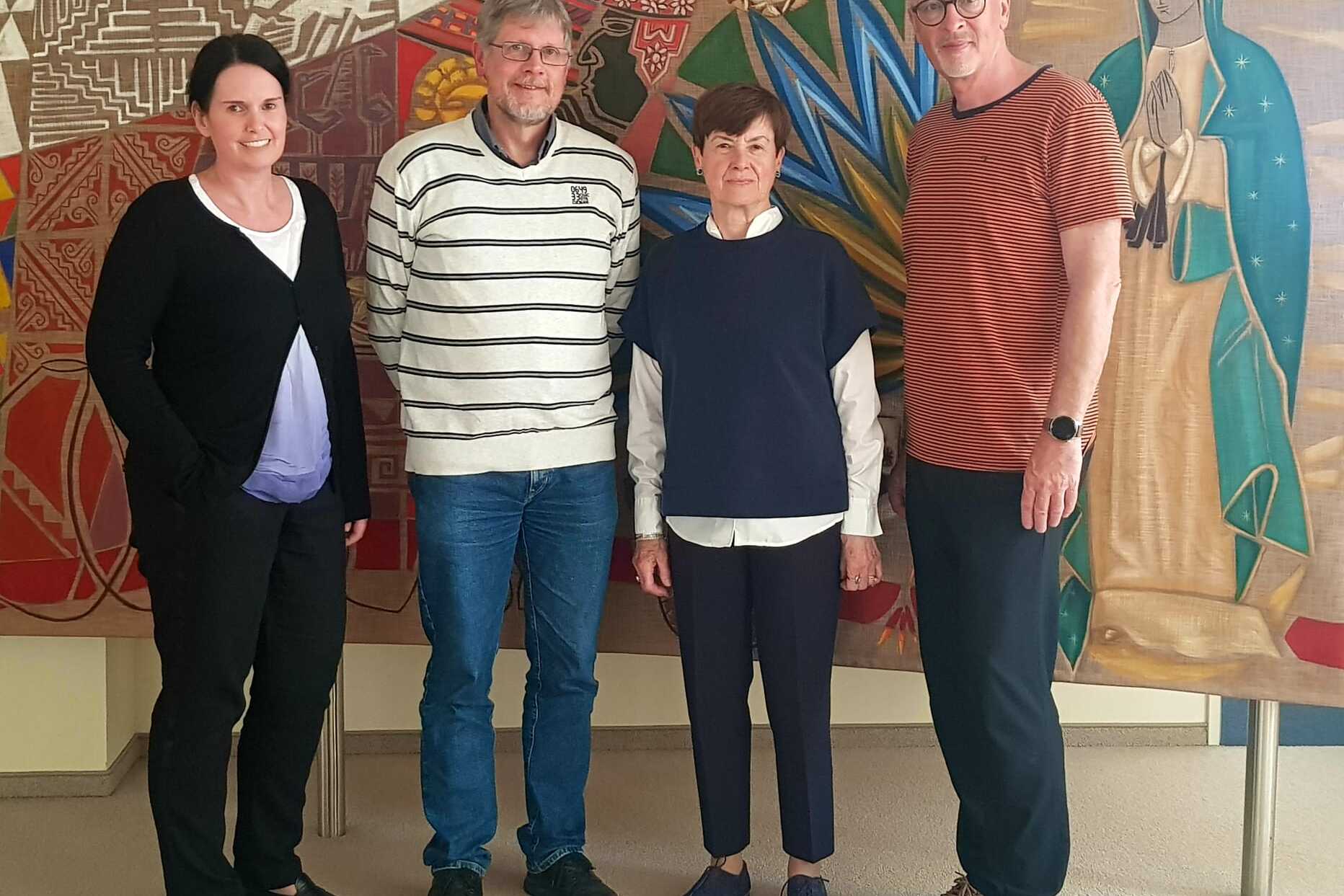 Vier Personen stehen vor einem bunten Wandgem&auml;lde in einem Innenraum.