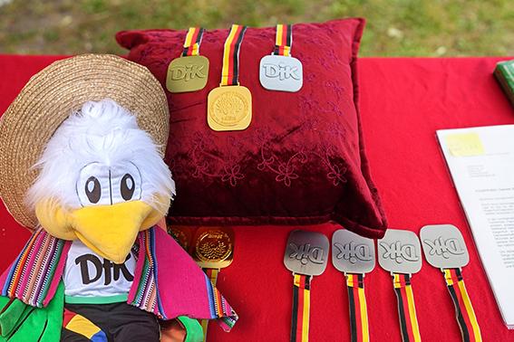 Pl&uuml;schfigur eines Vogels mit Strohhut und mehreren Medaillen auf rotem Tisch. Ein Kissen im Hintergrund.