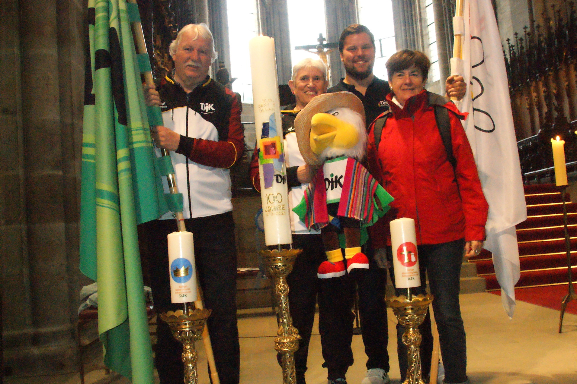 Vier Personen in Kirchenumgebung halten Fahnen und eine Maskottchenfigur zwischen zwei goldenen Kerzenhaltern.
