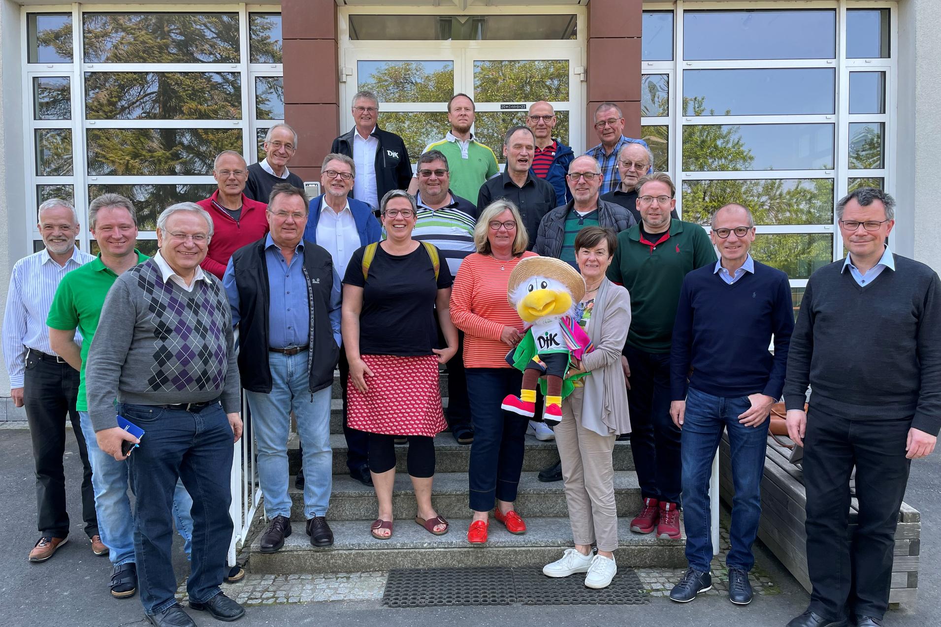 Gruppenfoto von 26 Personen vor einem Geb&auml;ude, darunter ein Maskottchen mit buntem Outfit.