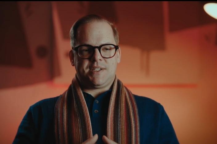 Mann mit Brille und gestreiftem Schal spricht vor einem hellen, unscharfen Hintergrund.