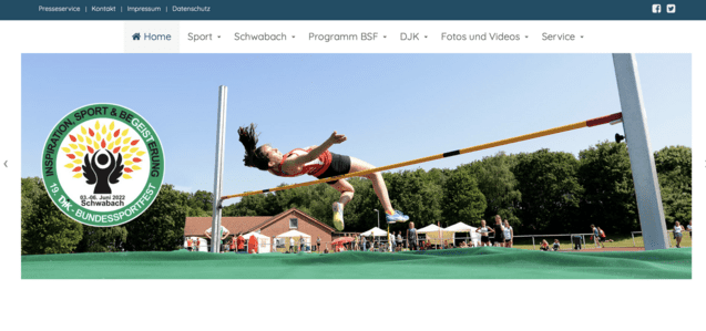 Weibliche Athletin springt &uuml;ber eine Hochsprunglatte auf einem Sportplatz, im Hintergrund stehen Zuschauer und Geb&auml;ude.