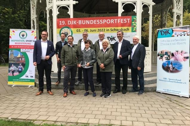 Gruppe von acht Personen posiert vor dem Pavillon mit Banner zum 19. DJK-Bundessportfest in Schwabach.