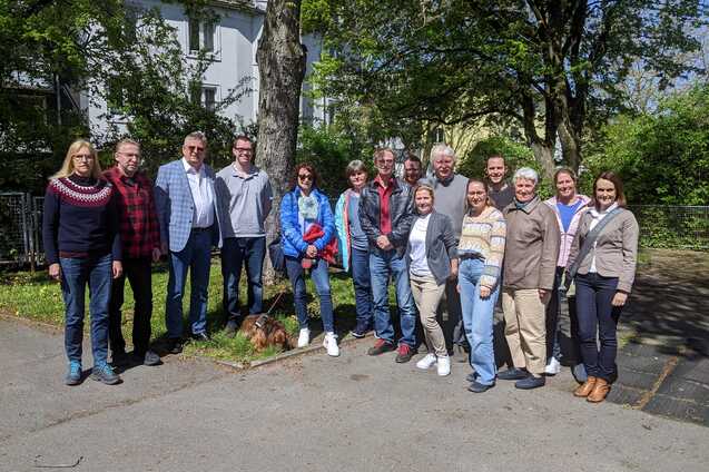 Gruppe von 14 Personen, stehend im Freien, umgeben von B&auml;umen, mit einem kleinen Hund im Vordergrund.