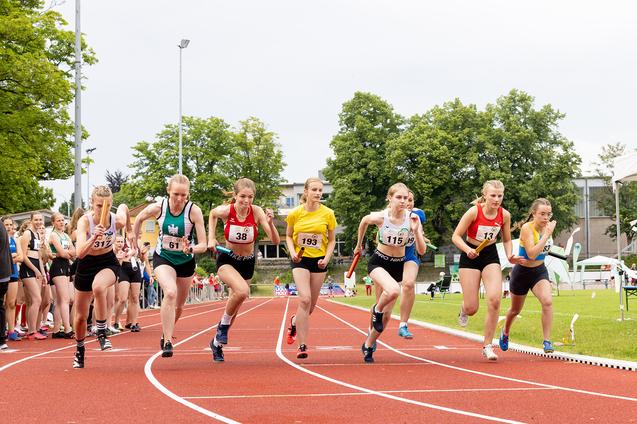 Sechs Athletinnen starten sprintend von der Startlinie auf einer Laufbahn, umgeben von gr&uuml;nem Gel&auml;nde.