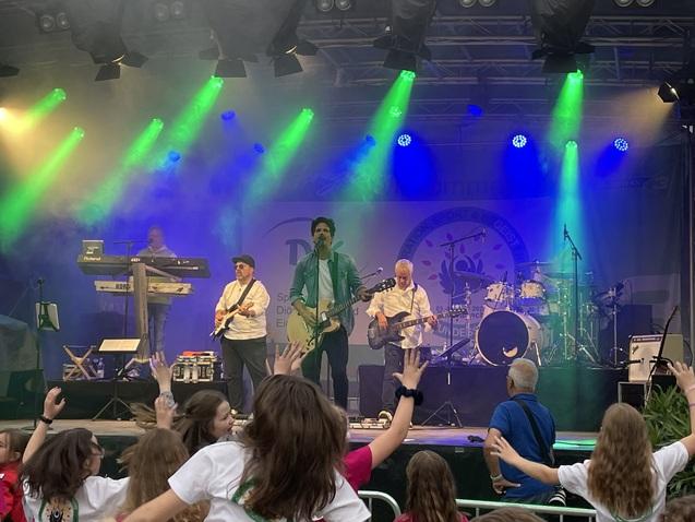 Musikband tritt auf einer Bühne mit bunten Lichtern auf, während das Publikum begeistert die Hände hebt.