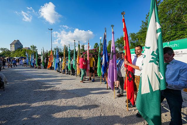 Menschen halten Fahnen in einer Reihe auf einem Platz, umgeben von Bäumen und einem blauen Himmel.