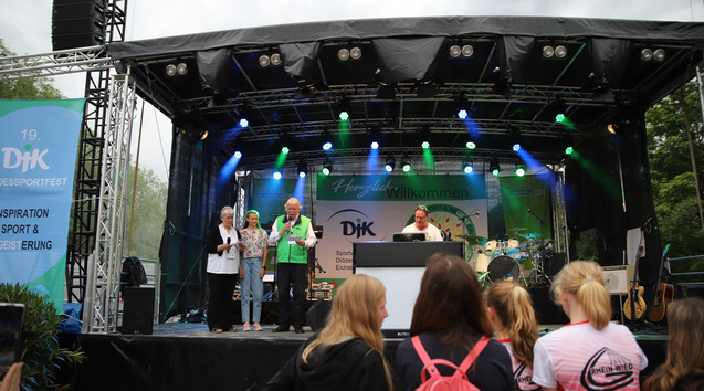 Auf einer B&uuml;hne stehen vier Personen vor einem Publikum, im Hintergrund sind Musikinstrumente und eine Begr&uuml;&szlig;ungsbanner.