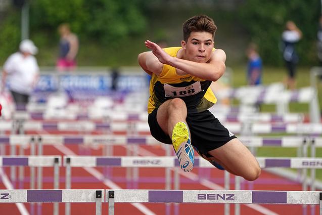 Ein junger Athlet springt über eine Hürde auf einer roten Tartanbahn während eines Wettkampfs.