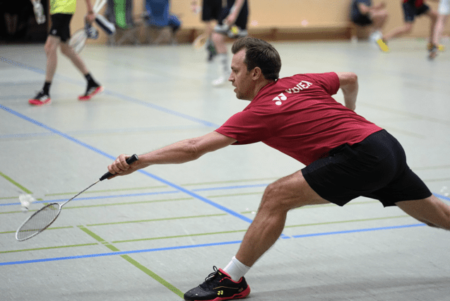 Mann in rotem Shirt spielt Badminton, während er sich in einer sportlichen Bewegung auf dem Hallenboden abstützt.