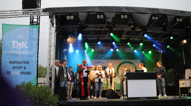 Veranstaltung auf einer B&uuml;hne mit mehreren Personen, die am Mikrofon stehen, umgeben von Lichtern und einem Banner.