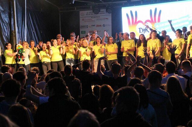 Gruppe von Menschen in gelben T-Shirts applaudiert auf einer B&uuml;hne vor einem Publikum, mit buntem Hintergrund.