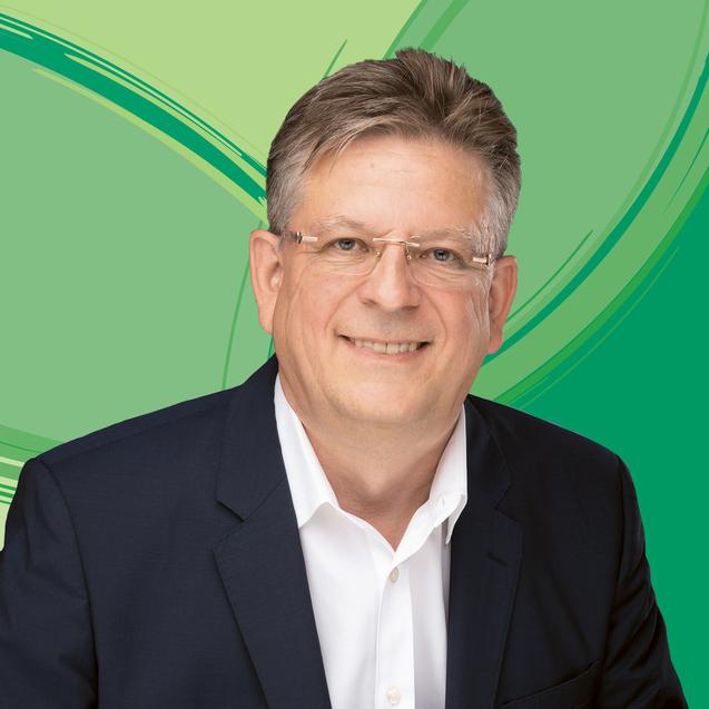 Portrait eines l&auml;chelnden Mannes in einem dunkelblauen Blazer vor einem gr&uuml;nen Hintergrund mit geschwungenen Linien.