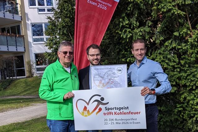 Drei M&auml;nner halten ein Plakat mit der Aufschrift &bdquo;Sportgeist trifft Kohlenfeuer&ldquo; vor einem Wohngeb&auml;ude und einem Fahnenmast.