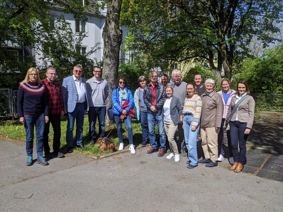 Gruppe von 14 Personen, stehend im Freien, umgeben von B&auml;umen, mit einem kleinen Hund im Vordergrund.