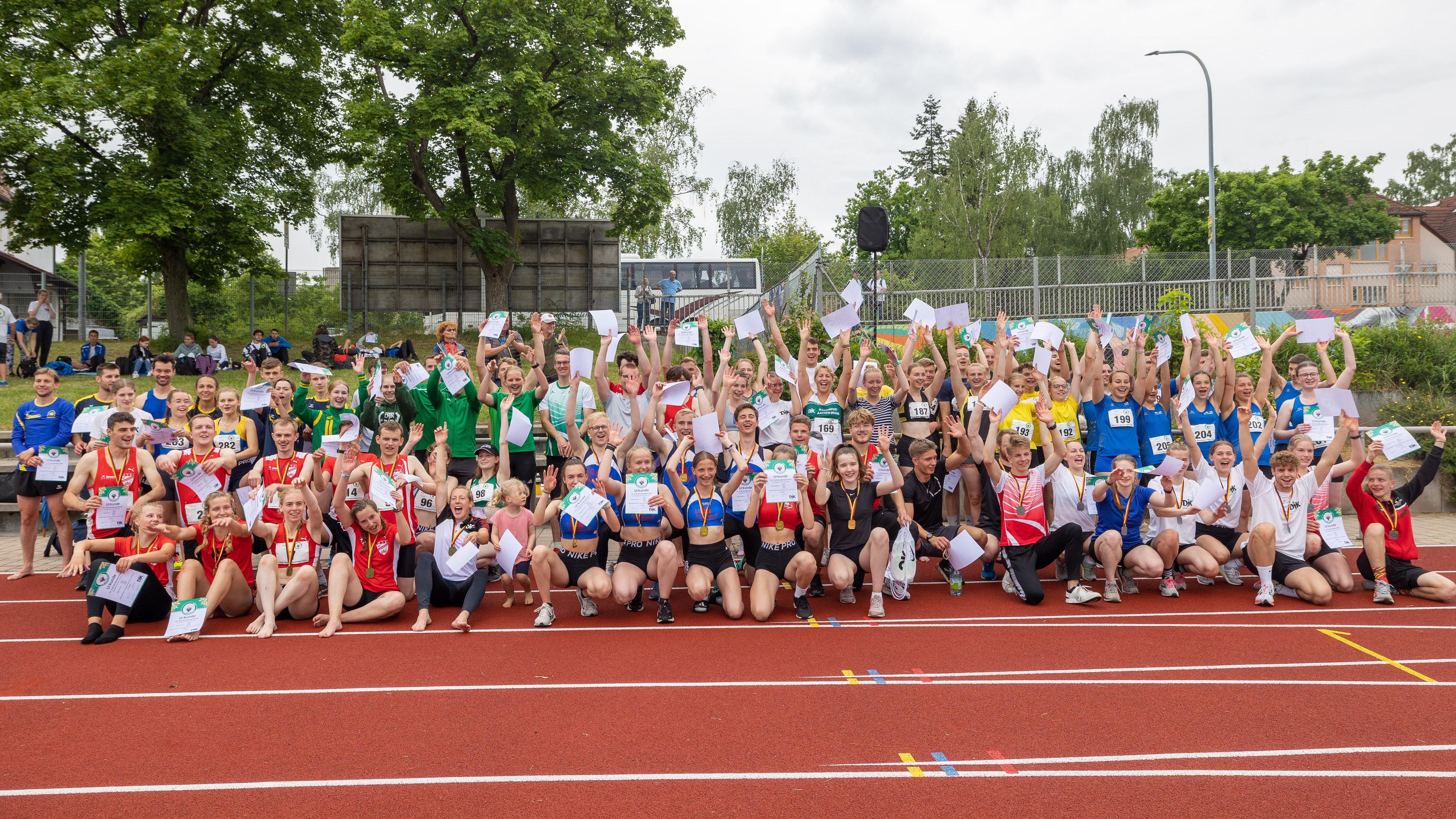 Gruppierung von Athleten in verschiedenen Trikots, die jubelnd auf einer Laufbahn stehen und kleine Papiere halten.