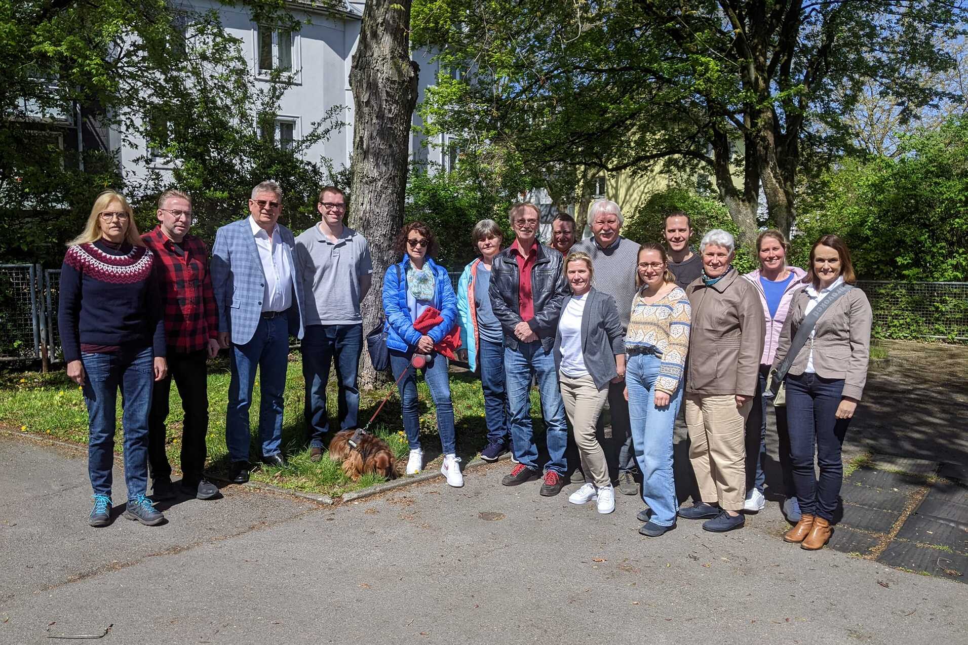 Gruppe von 14 Personen, stehend im Freien, umgeben von B&auml;umen, mit einem kleinen Hund im Vordergrund.