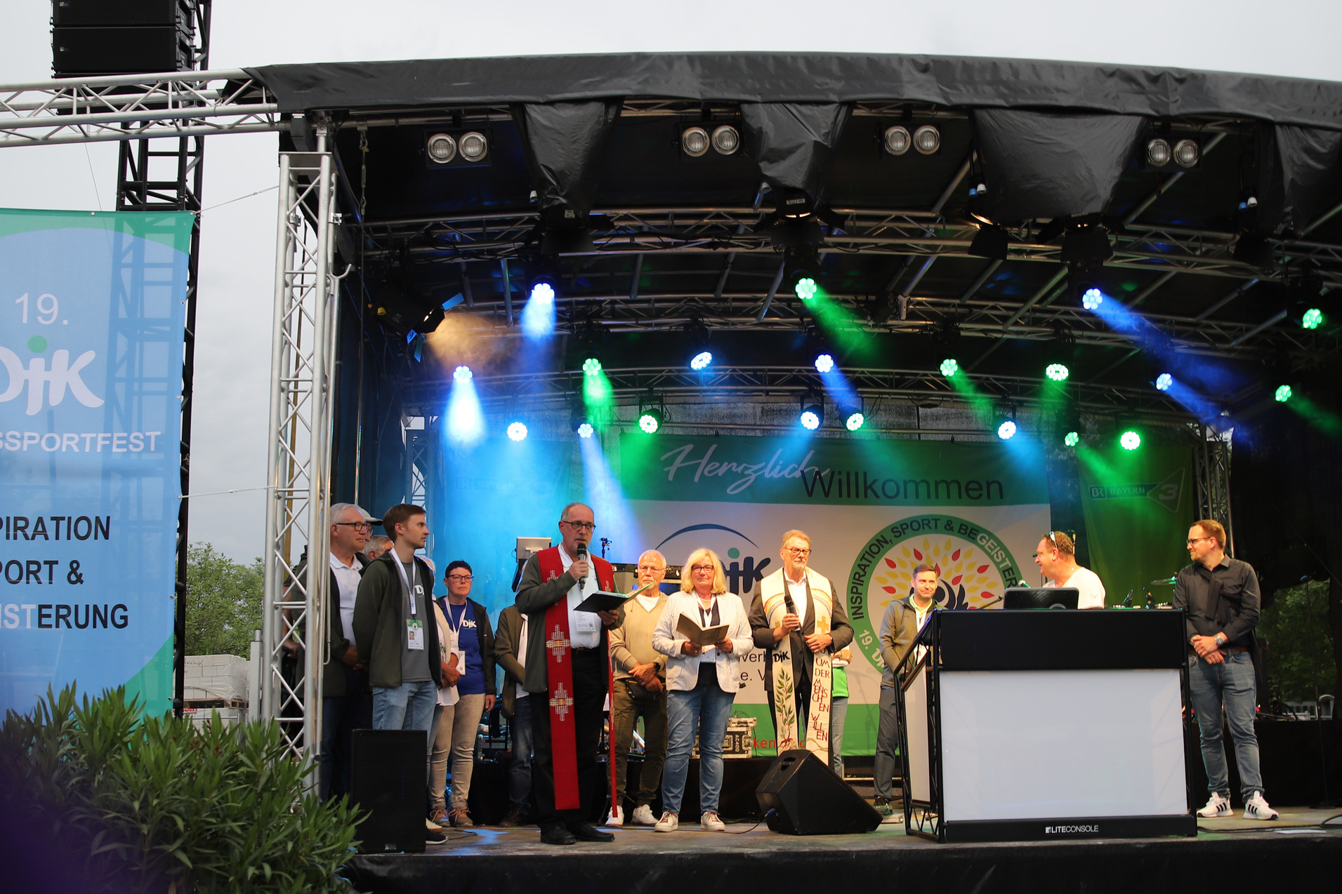 Veranstaltung auf einer B&uuml;hne mit mehreren Personen, die am Mikrofon stehen, umgeben von Lichtern und einem Banner.