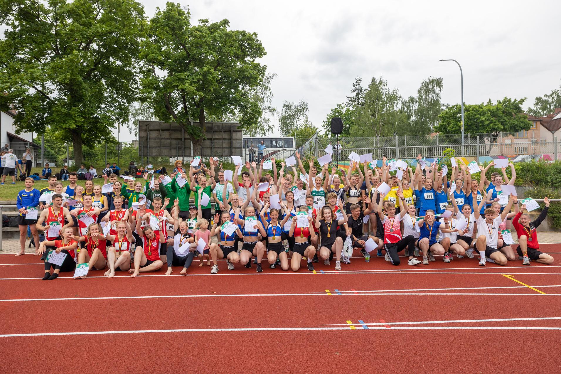 Gruppierung von Athleten in verschiedenen Trikots, die jubelnd auf einer Laufbahn stehen und kleine Papiere halten.