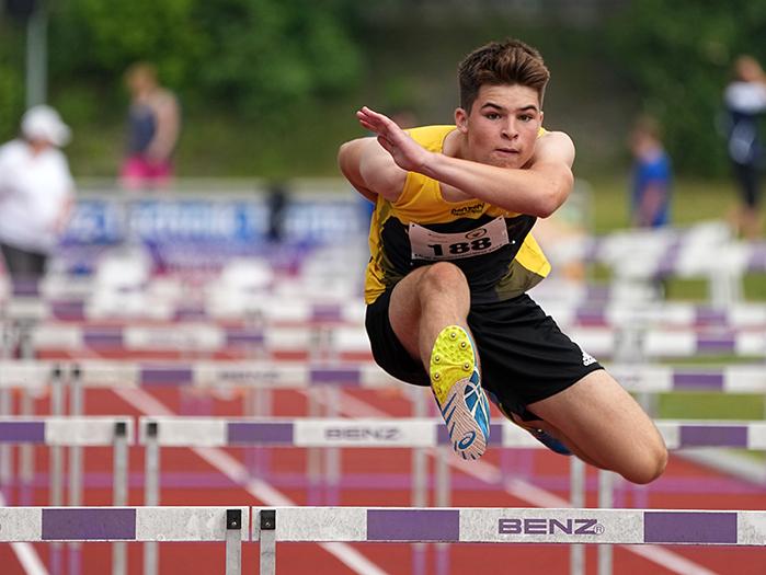 Ein junger Athlet springt über eine Hürde auf einer roten Tartanbahn während eines Wettkampfs.