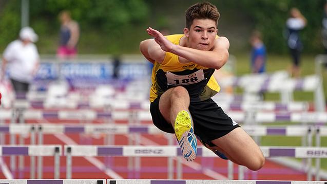 Ein junger Athlet springt &uuml;ber eine H&uuml;rde auf einer roten Tartanbahn w&auml;hrend eines Wettkampfs.