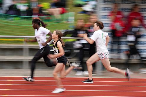 Drei junge Leichtathletinnen sprinten auf einer roten Laufbahn, Hintergrund mit Zuschauern und Sportausr&uuml;stung.