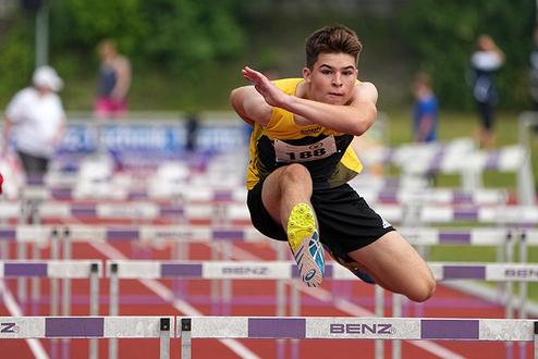 Ein junger Athlet springt &uuml;ber eine H&uuml;rde auf einer roten Tartanbahn w&auml;hrend eines Wettkampfs.