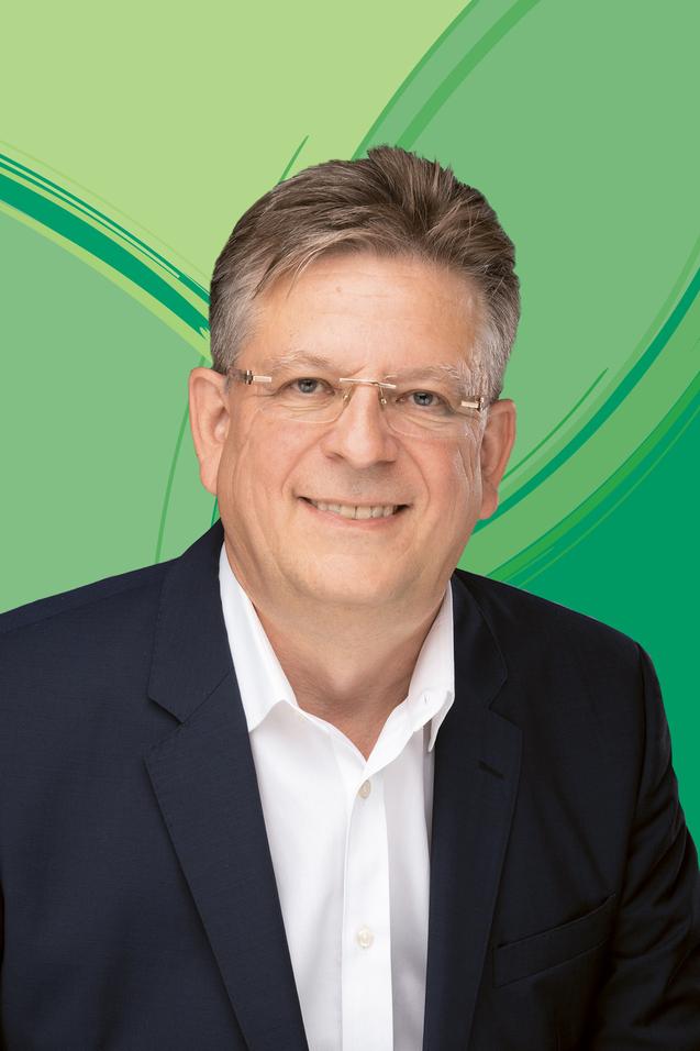 Portrait eines Mannes mit Brille in dunklem Anzug vor einem gr&uuml;nen, grafischen Hintergrund.