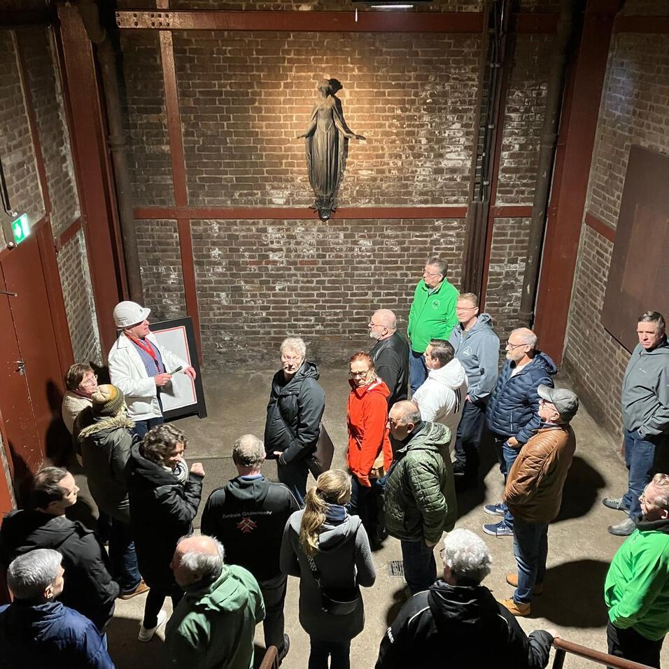 Gruppe von Menschen h&ouml;rt einem redenden Guide in einem alten, industrialisierten Raum mit einer Statue zu.