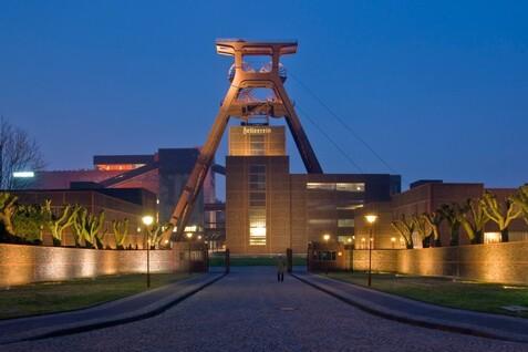 Eingang zu einem ehemaligen F&ouml;rderturm mit moderner Architektur und beleuchtetem Weg bei D&auml;mmerung.