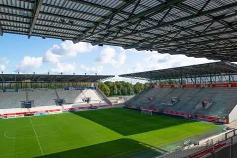 Blick auf ein leeres Fu&szlig;ballstadion mit gr&uuml;nem Rasen und Zuschauerr&auml;ngen unter blauem Himmel und Wolken.