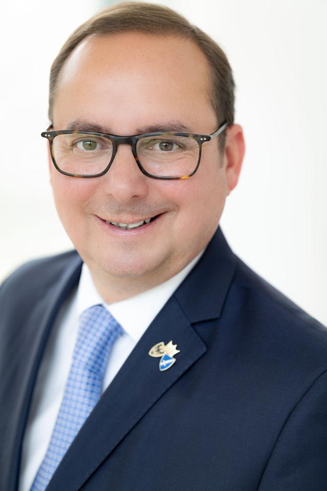 Portr&auml;t eines l&auml;chelnden Mannes mit Brille, Anzug und hellblauer Krawatte, mit Abzeichen auf der Brust.