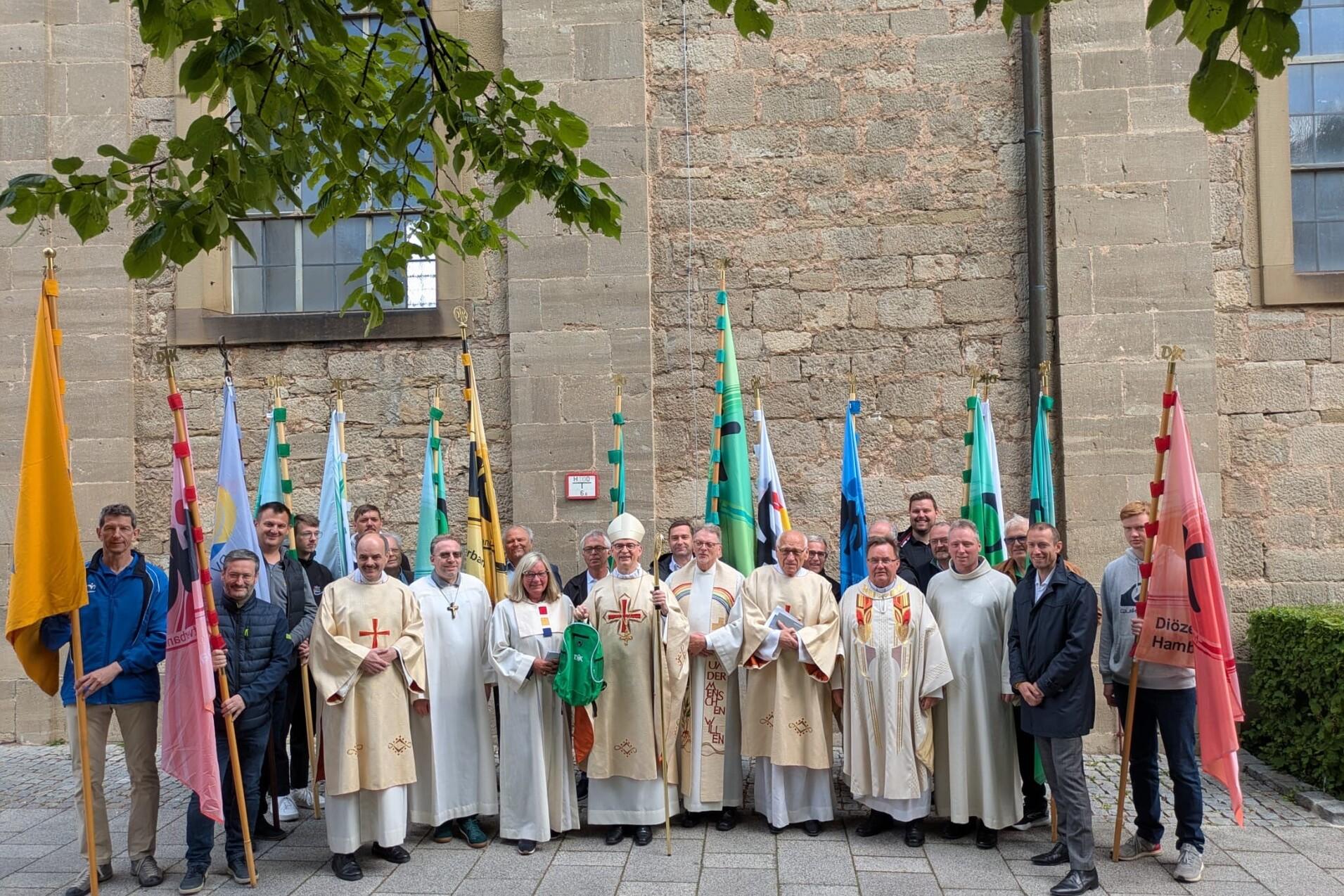 Gruppe von Geistlichen und Laien in religi&ouml;ser Kleidung mit Fahnen vor einer historischen Steinmauer.