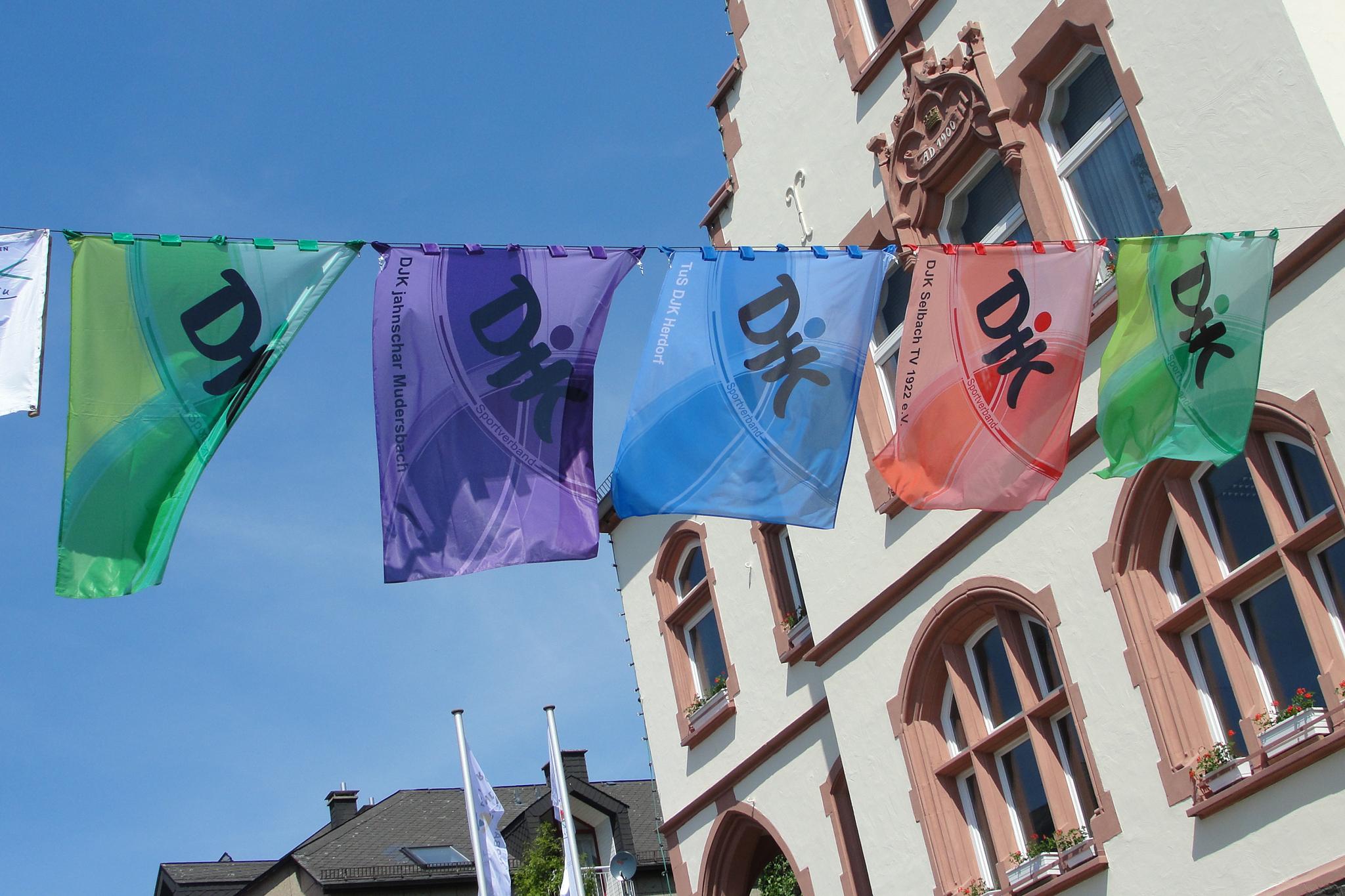 Farbenfrohe Fahnen mit Schriftzeichen h&auml;ngen zwischen historischen Geb&auml;uden in einem sonnigen Stadtbild.