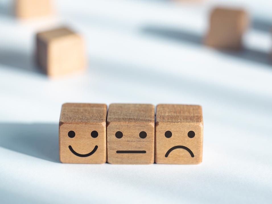 Drei Holzw&uuml;rfel mit Freude-, Neutral- und Trauer-Gesichtsausdr&uuml;cken auf einem hellen Hintergrund.