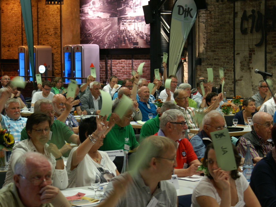 Gruppe von Menschen in einem Raum, die mit gr&uuml;nen Abstimmkarten ihre Stimmen erheben, w&auml;hrend sie an einer Veranstaltung teilnehmen.