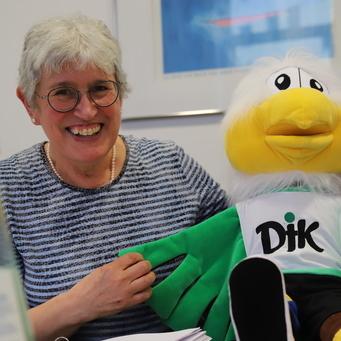 Zwei Personen l&auml;cheln und halten eine gro&szlig;e, gr&uuml;ne Maskottchenfigur mit gelbem Kopf und Ohr auf einem Tisch.