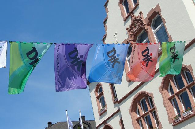 Farbenfrohe Fahnen mit Schriftzeichen h&auml;ngen zwischen historischen Geb&auml;uden in einem sonnigen Stadtbild.