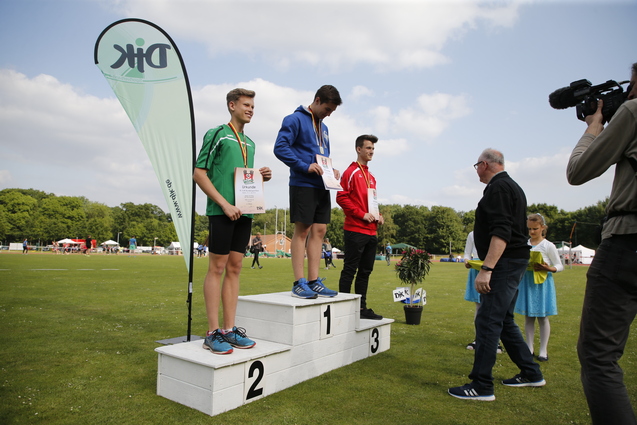 Drei Medaillengewinner stehen auf einem Podest, w&auml;hrend ein Zuschauer ihnen Urkunden &uuml;berreicht.