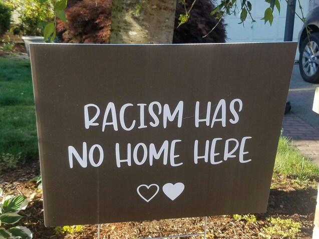 Schild mit der Aufschrift "RACISM HAS NO HOME HERE" in wei&szlig;en Buchstaben auf braunem Hintergrund, umgeben von Pflanzen.